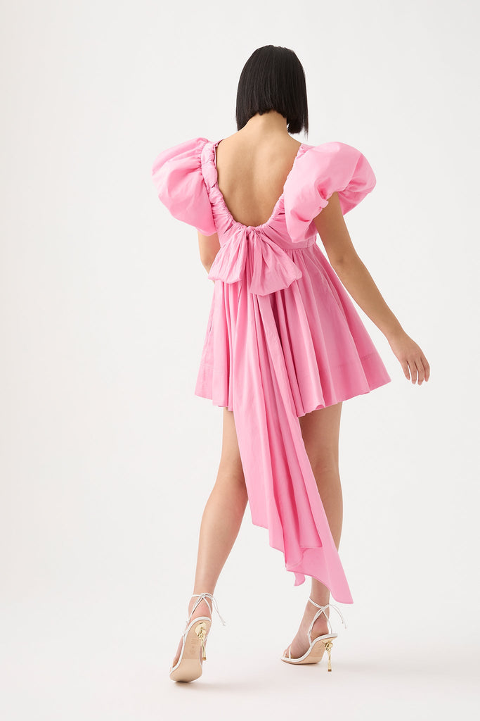 Woman turned back from the camera, wearing pink Gretta Bow Back Mini Dress from Aje featuring deep backline with bow and long ribbons adorned with short tulip sleeves.