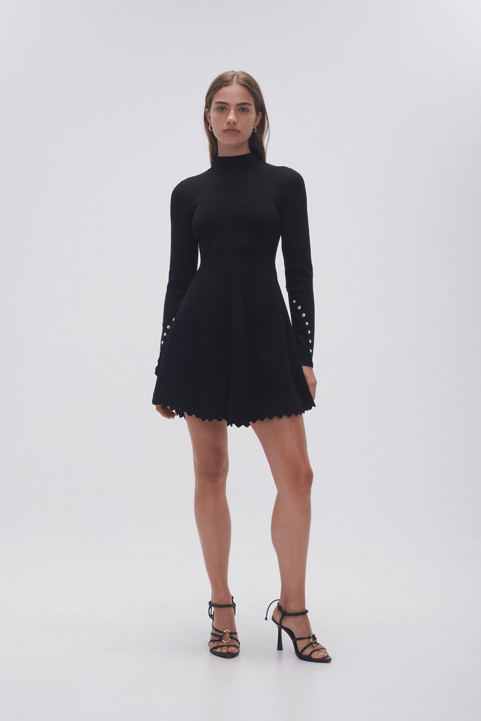 Woman wearing Aje Sylvia Knitted Mini Dress in black, featuring a turtleneck and long sleeves, adorned with delicate pearl beaded buttons.